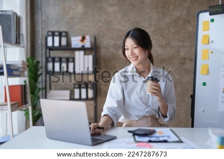 Happy Asian businesswoman working and checking work with laptop computer while having a comfortable cup of coffee at office. Royalty-Free Stock Photo #2247287673