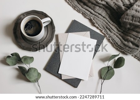 Moody breakfast still life, stationery. Blank greeting card, invitation mockup, dry eucalyptus tree branches, diary and linen blanket. Cup of coffee on white table background. Flat lay, top view. 