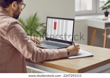 Concentrated male financier using laptop working in office with data entered in spreadsheet. Man fills out paper documents and enters data into electronic files marked in red on laptop screen. Royalty-Free Stock Photo #2247292111