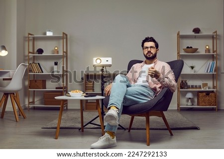 Relaxed young man sitting in a comfortable armchair in the living room at home, using a modern video projector, watching a movie, having a snack, and drinking tea