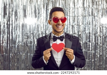 Portrait of funny handsome young dark-skinned man in suit with red heart in his hands on shiny silver background. Man in black suit and with heart-shaped glasses smiles with toothy smile at camera.