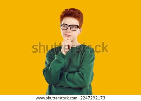 Thoughtful teenage boy standing with his hand on chin. Handsome puzzled redhead boy wearing glasses posing with serious face expression over yellow isolated background Royalty-Free Stock Photo #2247292173