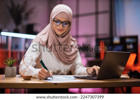 Focus in hand with pen. Attractive confident muslim business woman, office manager, wearing hijab using laptop while making financial report while writing on paper working at night.