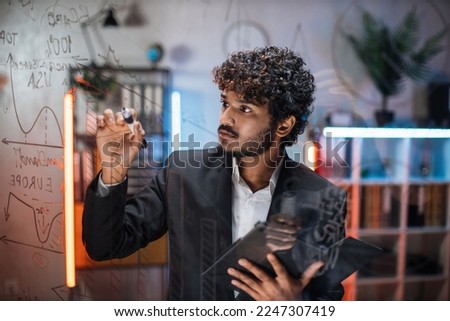Focused smart businessman, drawing graphs on glass presentation board. Male manager finance expert working in office at night, writing on glass board and using tablet computer