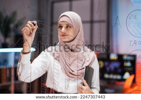 Confident female ceo manager, writing tasks or new startup ideas on glass wall, while checking project progress using tablet pc. Young businesswoman planning project on board in office.