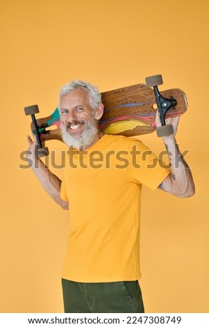 Smiling happy cool gray haired bearded hipster old senior man skater wearing t-shirt holding skateboard standing isolated on yellow background. Older people freedom spirit concept. Vertical