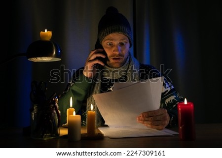 shocked man in candle light looking at high bills for electricity and gas heating in cold dark room at home. energy crisis, household expenses and inflation concept