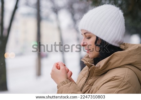 A young girl in a white hat warms her hands. It stands in the city in winter under the snow Royalty-Free Stock Photo #2247310803