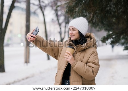 Cute girl laughing and making a video call outside in winter. Hold a disposable cup of coffee