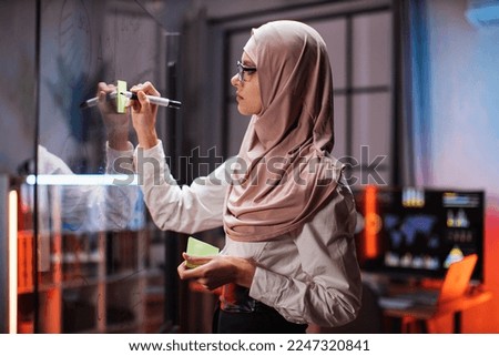 Focused muslim female employee writing on colorful sticky notes on office glass board, developing business project, using tablet pc. Startup plan, people and technologies concept.