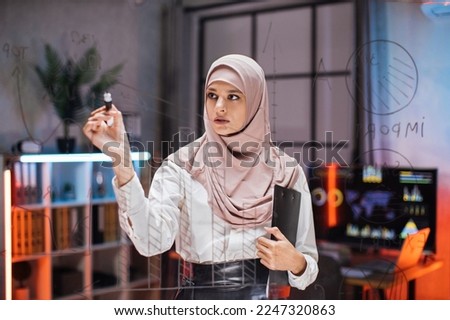 Confident female ceo manager, writing tasks or new startup ideas on glass wall, while checking project progress using tablet pc. Young businesswoman planning project on board in office.