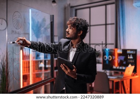 Confident male ceo manager, writing tasks or new startup ideas on glass wall, while checking project progress using tablet pc. Confident businessman planning project on board in office