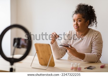Disappointed beauty blogger curly hispanic overweight young lady sitting at vanity table at home, showing eyeshadow palette at phone camera, influencer making review, dont recommend beauty product