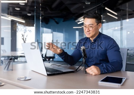 A young Asian man is sitting at a desk in the office. Speaks into the microphone through a video call from a laptop. Online business meeting, blogger, recording interviews, podcast.