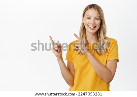 Portrait of young woman demonstrating, showing announcement or logo, pointing fingers left, standing in yellow t-shirt over white background