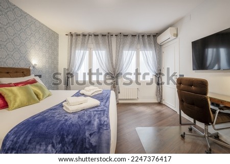 Bedroom with a headboard upholstered in brown velvet in a bedroom with satin cushions of various colors, windows with translucent curtains, a desk, a TV on the wall and a wallpapered wall
