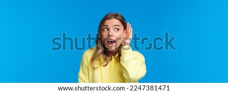 Close-up portrait of intrigued and curious blond young girl with pierced ear, gossiping, eavesdropping and listening to someone conversation interested, standing blue background. Copy space Royalty-Free Stock Photo #2247381471