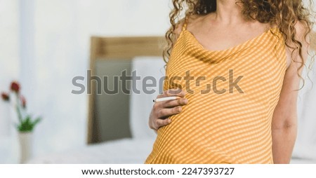 Curly hairstyle young unhappy unhealthy bad behavior Caucasian pregnancy mother in maternity long dress cloth sit on bed in bedroom holding smoking cigarette taking risk and danger to unborn child.