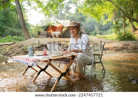 Asian woman travel and camping alone reading a book and using laptop while relaxing on the deck chairs in the river at natural park in Thailand. Recreation and journey outdoor activity lifestyle.