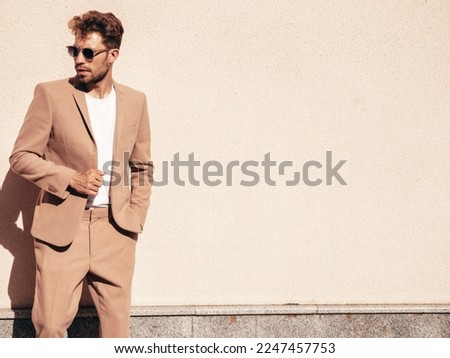 Handsome confident stylish hipster lambersexual model.Sexy modern man dressed in elegant beige suit. Fashion male posing in the street in city at sunset. In sunglasses. Near wall Royalty-Free Stock Photo #2247457753