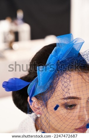 Cropped close-up shot of a young woman with a dark blue fascinator with a bow and a veil decorated with feathers. A girl in a white blouse and a fascinator is posing in the studio. Side view.          Royalty-Free Stock Photo #2247472687