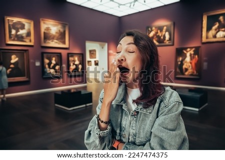 a bored girl visitor or student in an art gallery or museum yawns and looks at the masterpieces of classical artists