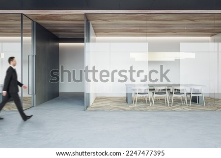 Businessman walking on concrete floor by stylish conference rooms with minimalistic style furniture on wooden parquet floor behind glass partitions in spacious office hall Royalty-Free Stock Photo #2247477395