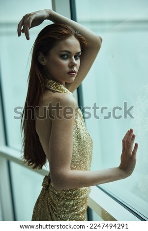 Beautiful fashion woman in stylish expensive gold sequined dress poses looking out the window at the city in a hotel, Christmas party