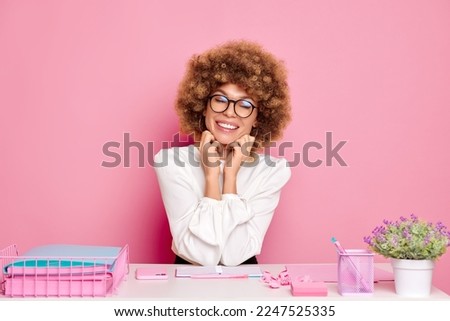A shot of pretty business woman, sitting at the office next to the pink wall, has toothy smile, works on the project, surrounded with documents and office goods, isolated over pink background