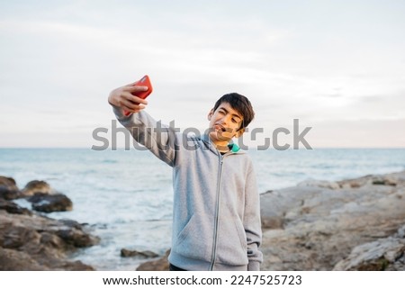 Boy taking a selfie on a red cellphone, on a rock breakwater, by the sea