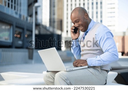 Stylish African American businessman talking on the phone while using a laptop computer outdoors. Royalty-Free Stock Photo #2247530221