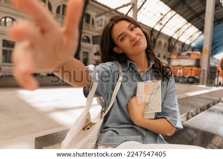 Cheerful young caucasian woman looking streching hand into camera sitts on platform with map. Model wears jeans jacket and shopper. Selfie, adventure concept.