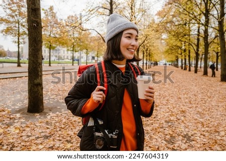 Beautiful smiling young asian woman wearing warm clothes drinking takeaway coffee during walk in autumn park