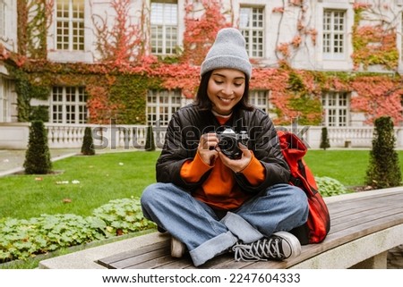 Beautiful young smiling asian woman in warm clothes taking pictures with vintage camera while sitting on bench in old city