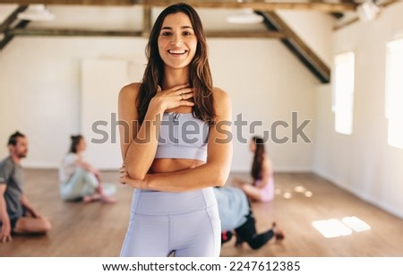 Sporty young woman smiling at the camera while standing in a yoga studio with a group of people in the background. Happy young woman attending a yoga class in a community fitness studio. Royalty-Free Stock Photo #2247612385