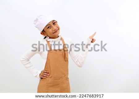 Cheerful little girl in chef uniform presenting something at blank space. Isolated on white background