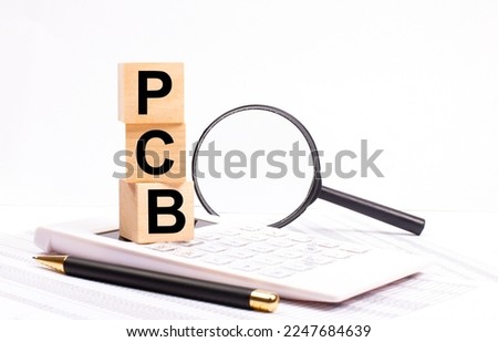 On a white calculator, next to a magnifying glass and a black pen, wooden cubes marked PCB Printed Circuit Board.