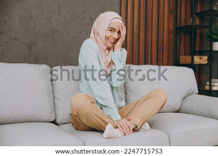 Full body shy calm fun young muslim woman wear hijab casual clothes sits on sofa couch stay at home flat rest relax spend free spare time in living room indoor. People uae middle eastern islam concept Royalty-Free Stock Photo #2247715753