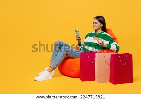 Full body happy young latin woman wear green knitted sweater sit in bag chair near package bags after shopping use mobile cell phone isolated on plain yellow background Black Friday sale buy concept Royalty-Free Stock Photo #2247715821