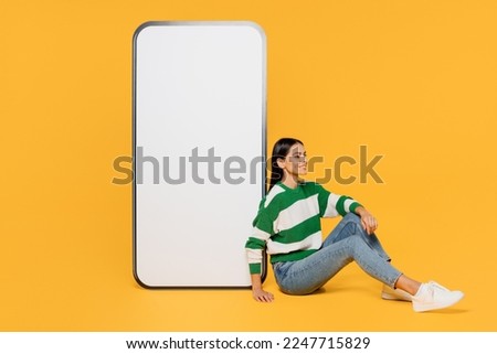Full body side view fun young latin woman wear casual cozy green knitted sweater near big huge blank screen mobile cell phone smartphone with area isolated on plain yellow background studio portrait