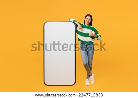Full body young happy latin woman wear casual cozy green knitted sweater big huge blank screen mobile cell phone smartphone with area show thumb up isolated on plain yellow background studio portrait