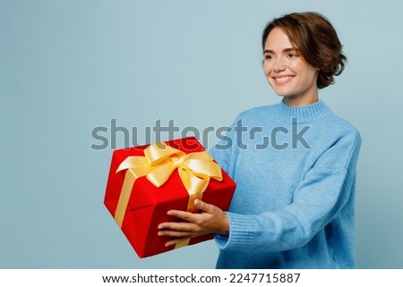 Side view young fun woman wear knitted sweater hold in hand giving red present box with gift ribbon bow isolated on plain pastel light blue cyan background studio portrait. People lifestyle concept