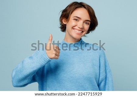 Young satisfied happy fun caucasian woman wear knitted sweater look camera showing thumb up like gesture isolated on plain pastel light blue cyan background studio portrait. People lifestyle concept