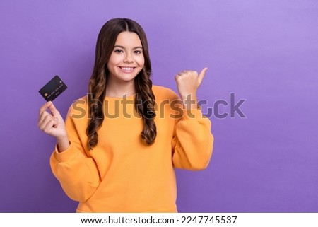 Photo of cheerful cute girl dressed orange sweatshirt rising bank card thumb empty space isolated purple color background