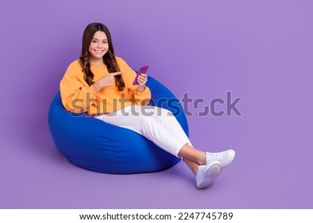Full length photo of sweet positive girl dressed orange sweatshirt bean bag pointing device empty space isolated purple color background