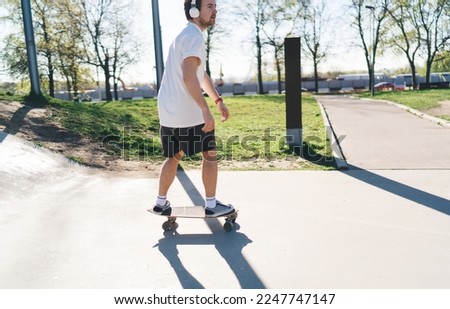 Side view of male skateboarder in summer sportswear riding skateboard at skate park while practicing stunt and enjoying music on headphones