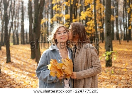Mother kiss in cheek teenage daughter in sunny golden forest. Happy girls hold bouquet of fallen yellow maple leaves and walk in autumn park. Beautiful fall nature, family lifestyle.