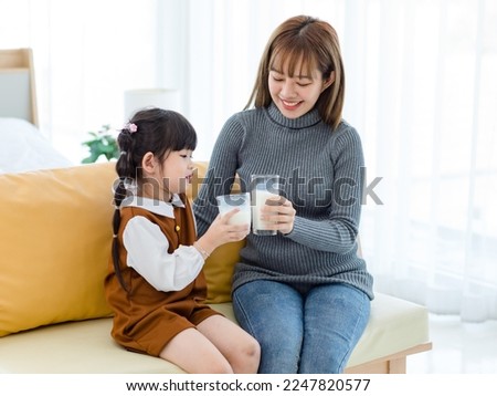 Millennial Asian young pretty female teenager mother nanny babysitter in casual outfit sitting on sofa smiling holding serving delicious milk glass to little cute preschooler daughter girl drinking. Royalty-Free Stock Photo #2247820577