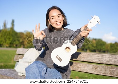Cute smiling girl shows ok sign and her new ukulele, sits on bench in park, recommends musical instrument.