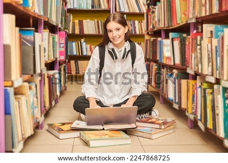 Educational concept. A modern girl student in a white shirt and black jeans sits on the floor in a library reading joyfully a book. Modern generation. Reading.